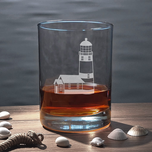 Glass with lighthouse design filled with amber liquid on a wooden surface with shells.