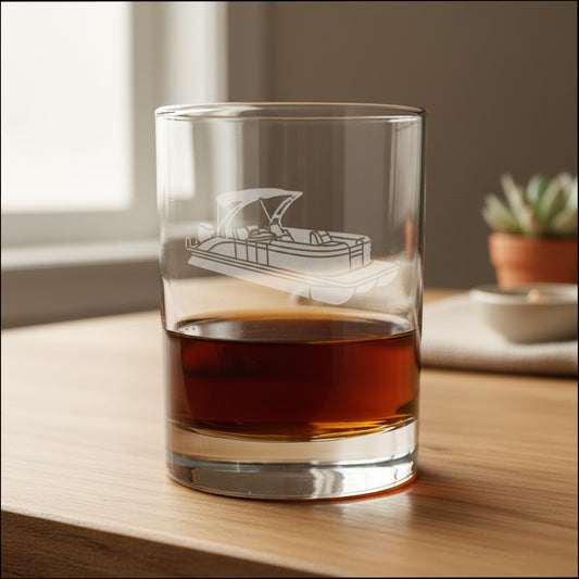 A clear rocks glass with an etched boat design, partially filled with amber liquor, sitting on a wooden table in a softly lit indoor setting.