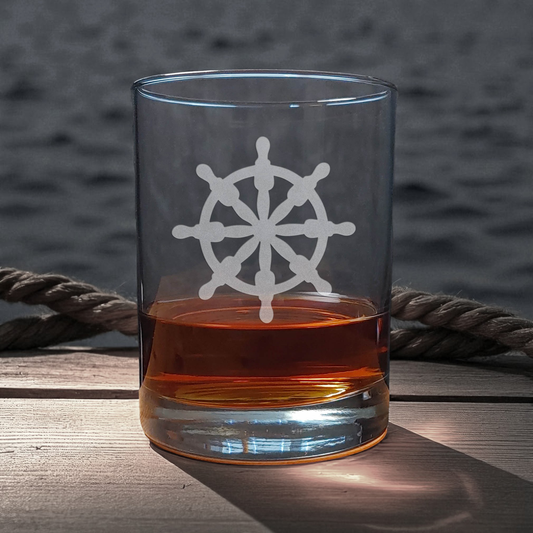 Clear glass with a white ship's wheel design filled with amber liquid on a wooden surface with a blurred water background.