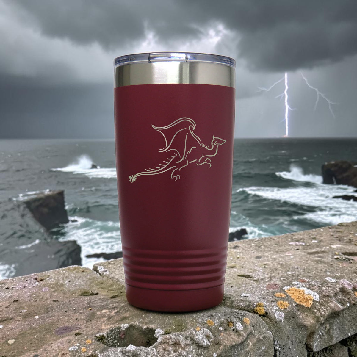 A dark red insulated travel tumbler with a silver stainless steel rim and a laser-engraved dragon design, set against a dramatic stormy ocean background with lightning.