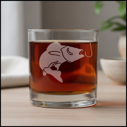 A clear lowball glass tumbler with a frosted pickerel and hook design, filled with amber liquid, set on a light wood surface with a softly blurred indoor background.