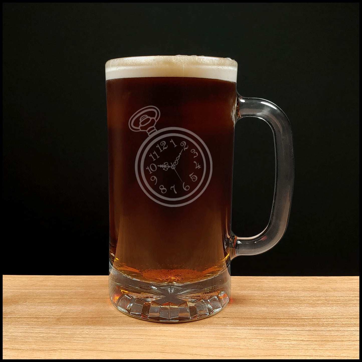 A clear glass beer mug with a handle, filled with dark amber beer and topped with white foam, featuring an etched pocket watch design on the front, set on a light wood surface against a black background.