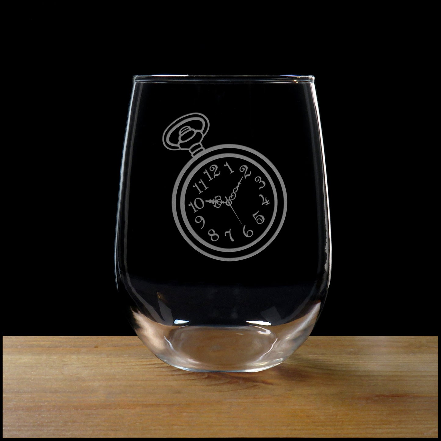 A clear stemless wine glass with an etched pocket watch design on the front, resting on a wooden surface against a solid black background.