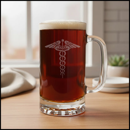 A clear glass beer mug filled with amber beer, standing on a wooden table in a softly blurred kitchen setting. The mug has an etched medical-style caduceus symbol printed on the front.