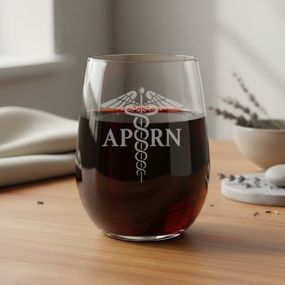 A stemless wine glass filled with red wine sits on a wooden table. The glass is etched with a caduceus symbol and  'APRN'.