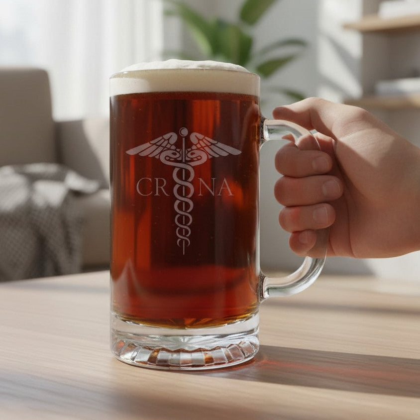 A hand holds a large clear glass beer mug filled with amber beer and topped with white foam. The mug has a frosted white medical-style emblem and the name 'CRNA' printed on the front, set on a wooden table in a softly blurred indoor room.