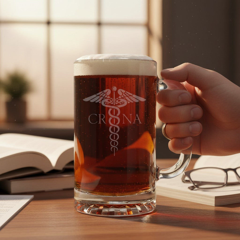 A hand holds a frosted glass beer mug filled with amber beer, featuring a white medical caduceus symbol and the engraved name 'CRNA'. The mug sits on a wooden desk with books and eyeglasses in a warm, sunlit room.