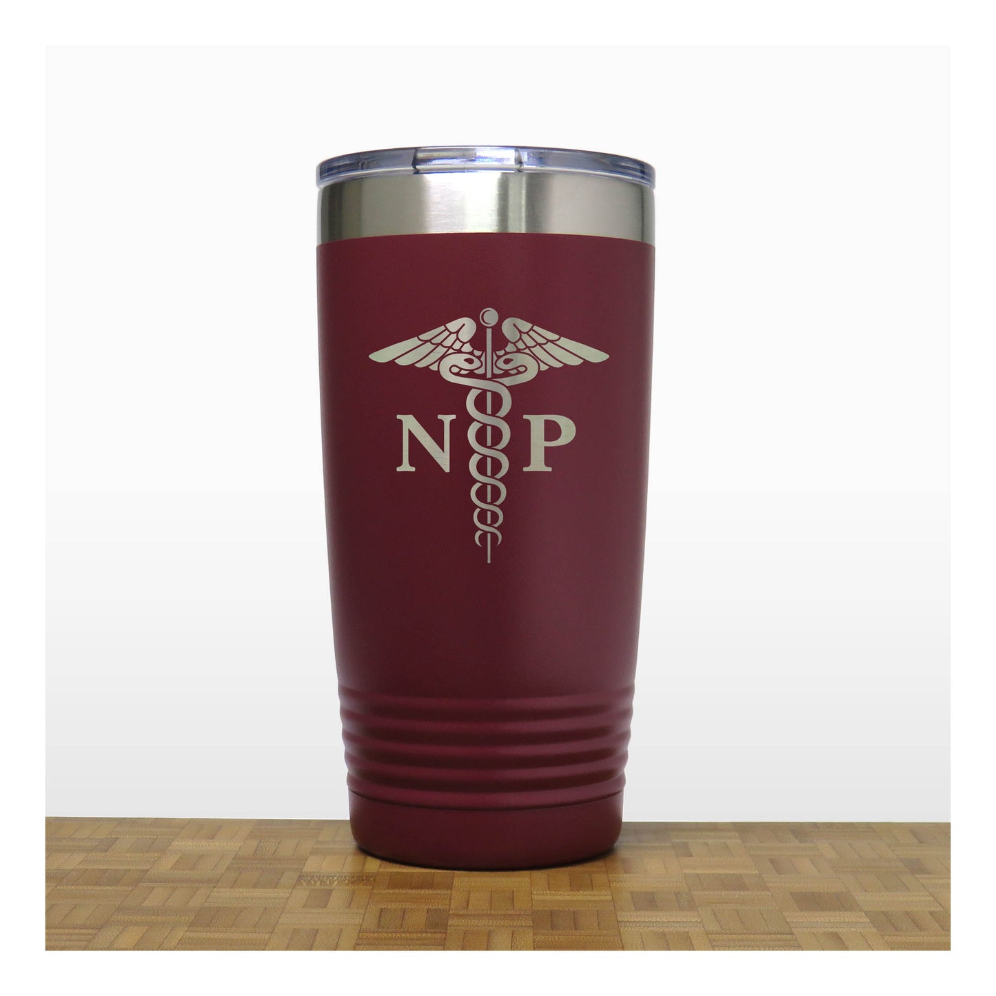 A maroon insulated travel tumbler with a stainless steel rim and a clear lid, with an laser engraved  medical caduceus symbol with the letters 'NP' on the front. The tumbler is shown standing on a wooden surface against a white background.