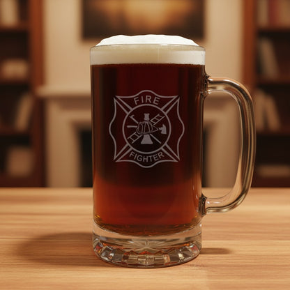A clear glass beer mug filled with amber beer and topped with white foam, standing on a wooden table. The mug has an etched Maltese cross firefighter emblem with the words 'FIRE FIGHTER' in the center.