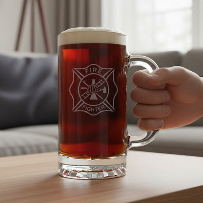 A clear glass beer mug with a handle, filled with amber beer and topped with foam, standing on a wooden table in a living room. On the front of the glass is an etched Maltese cross firefighter emblem