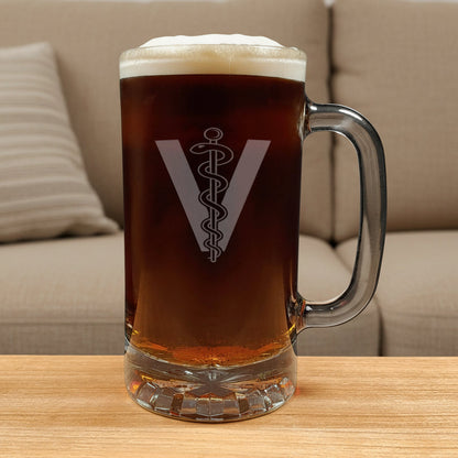 A clear glass beer mug filled with dark beer and topped with white foam, featuring a Veterinarian Symbol comprising of a "V", there is a rod in the middle with a snake wrapped around it , set on a wooden surface against a beige couch