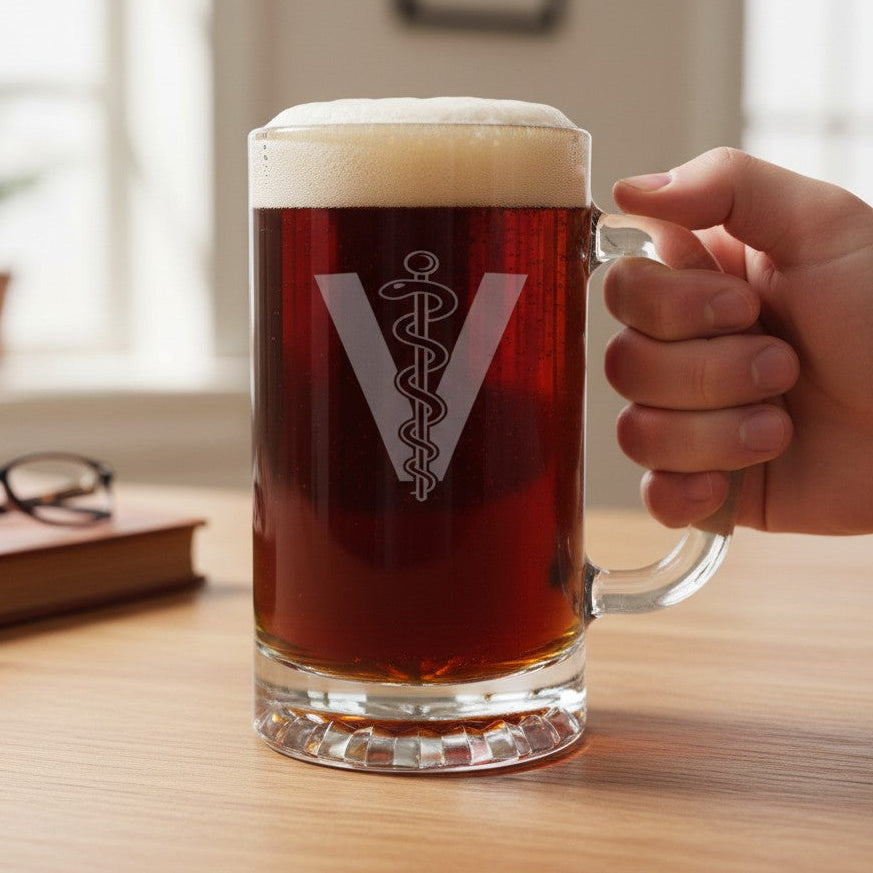 A hand holds a clear glass beer mug filled with dark amber beer and topped with white foam, featuring a Veterinarian Symbol comprising of a "V" with a rod with a snake wrapped around it in the middle, set on a wooden surface against a beige couch