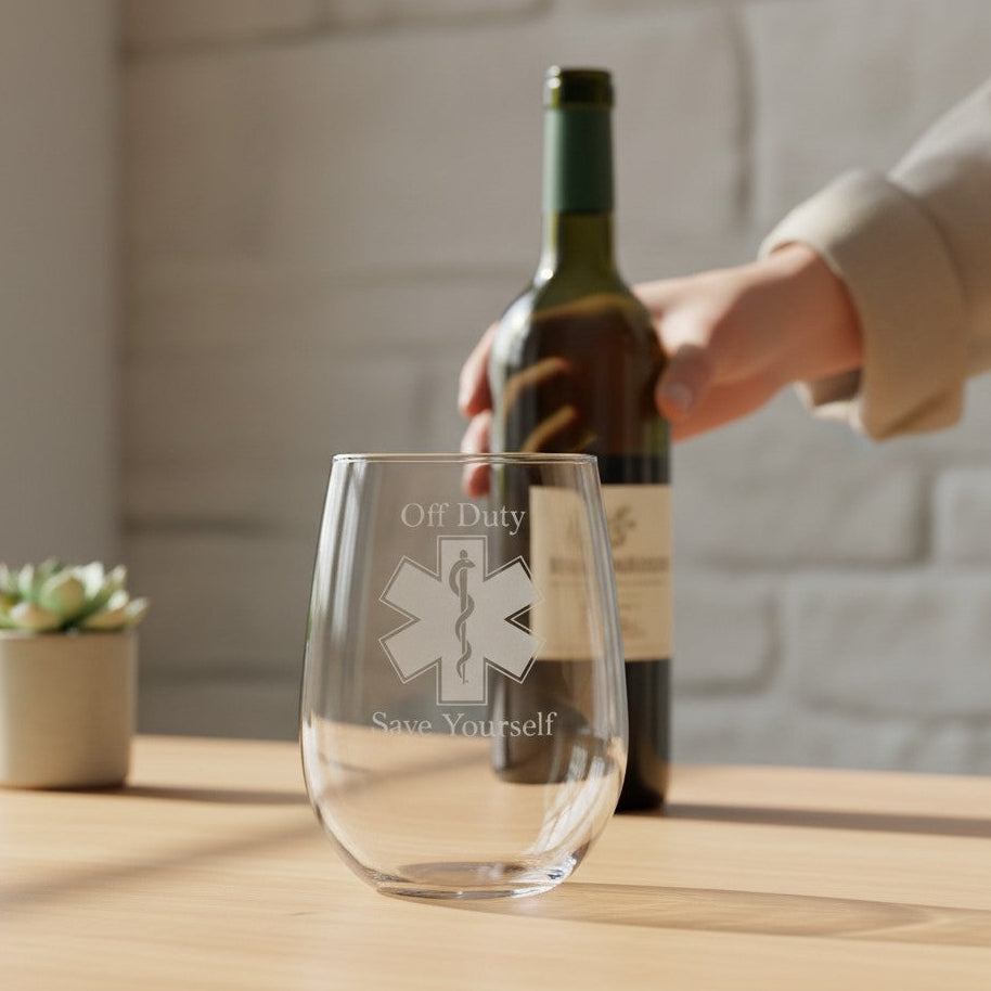A clear stemless wine glass with an etched EMS symbol and the words 'Off Duty Save Yourself' sits on a wooden table in a softly lit room. A hand holding a wine bottle is blurred in the background, with a small potted succulent on the left.