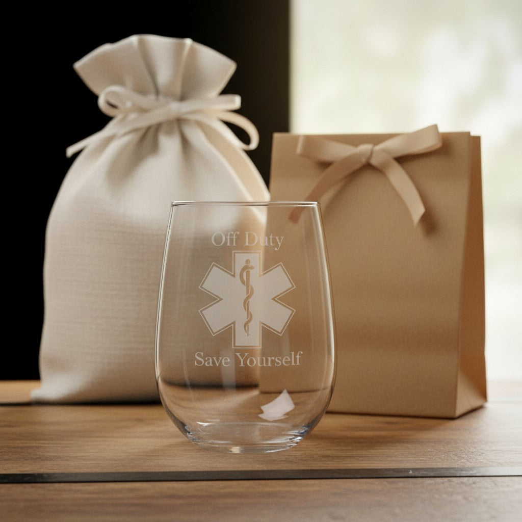 A clear stemless wine glass with etched text reading 'Off Duty Save Yourself' and a Star of Life medical symbol, displayed on a wooden surface with a cream drawstring pouch and a brown gift bag in the background.