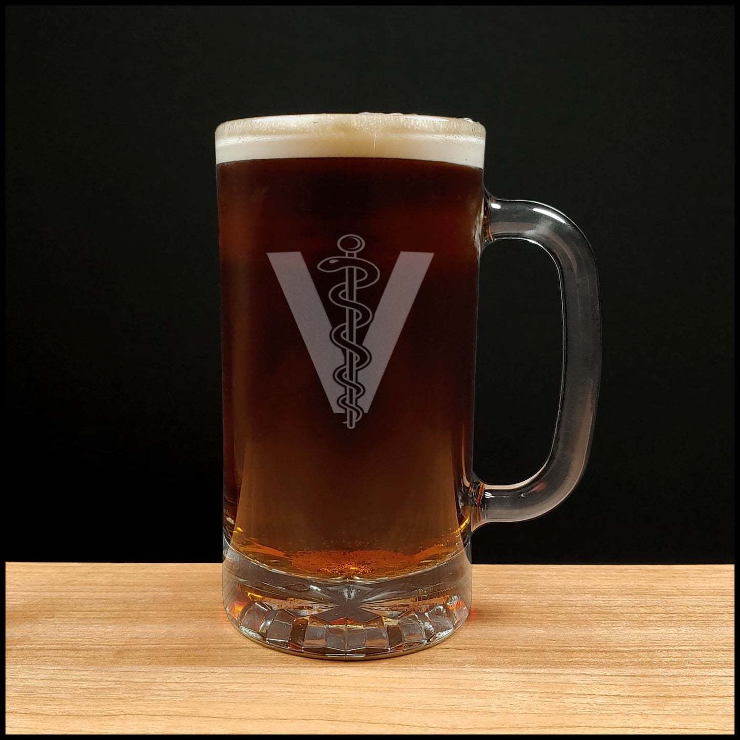 A clear glass beer mug filled with dark beer and topped with white foam,featuring a Veterinarian Symbol comprising of a "V", there is a rod in the middle with a snake wrapped around it,  set on a wooden surface against a black background.
