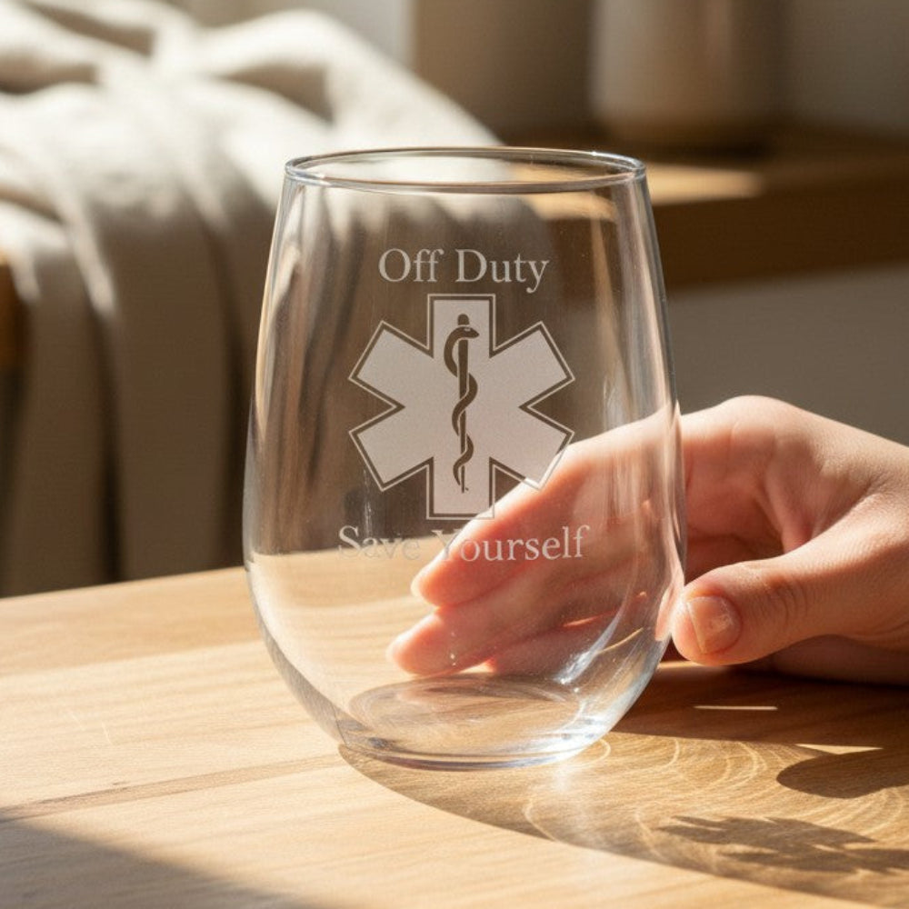 A clear stemless wine glass with an etched EMS symbol and the words 'Off Duty' and 'Stay Yourself' sits on a wooden table, with a person's hand resting near it in a softly lit indoor setting.