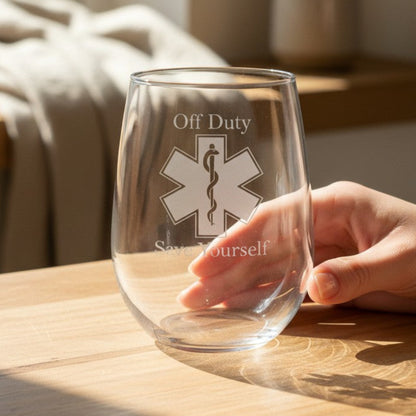 A clear stemless wine glass with an etched EMS symbol and the words 'Off Duty' and 'Stay Yourself' sits on a wooden table, with a person's hand resting near it in a softly lit indoor setting.