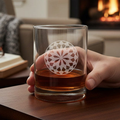 A hand holds a clear glass tumbler with a white geometric mandala design over an amber drink, set on a wooden table in a cozy living room with a fireplace in the background.