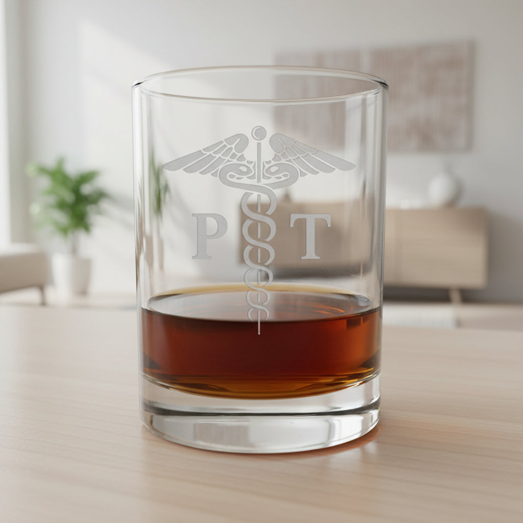 A clear glass rocks tumbler with an etched medical caduceus and the letters 'P T' on the front, partially filled with amber whiskey, set on a wooden surface against a dark gray textured background.