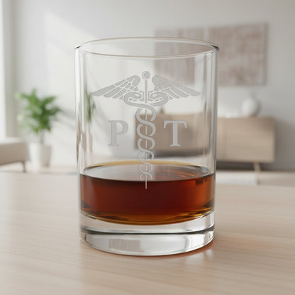 A clear glass rocks tumbler with an etched medical caduceus and the letters 'P T' on the front, partially filled with amber whiskey, set on a wooden surface against a dark gray textured background.