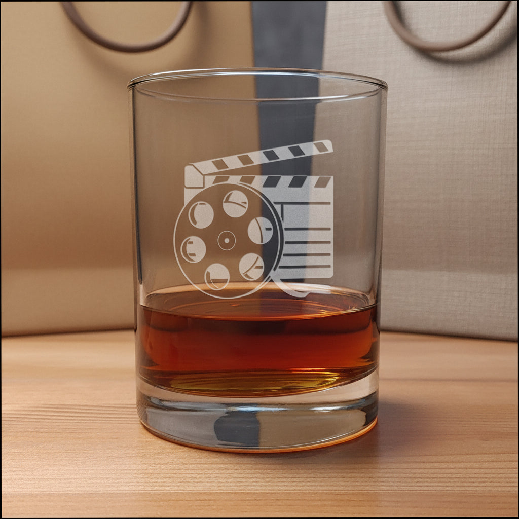 A clear glass rocks tumbler with an etched film clapperboard and reel design, partially filled with amber whiskey, set on a wooden surface with gift bags in the background.