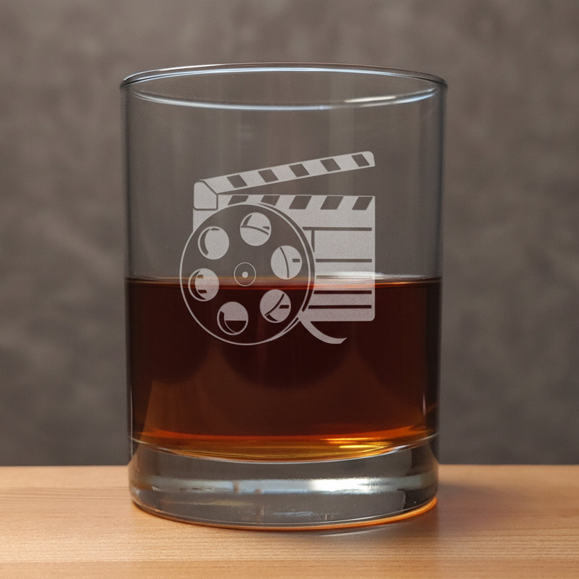 A clear glass rocks tumbler with an etched film clapperboard and reel design, partially filled with amber whiskey, set on a wooden surface against a  textured background.