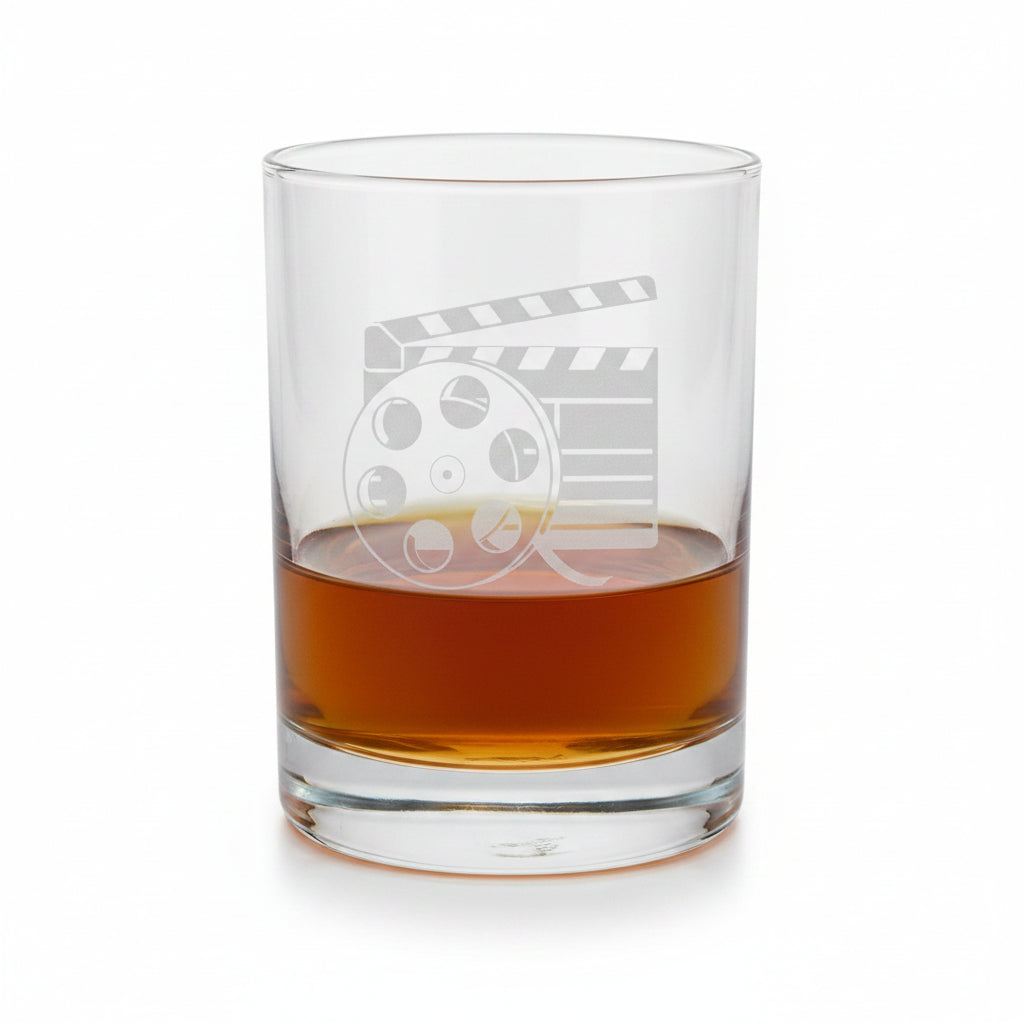 A clear glass rocks tumbler with an etched film clapperboard and reel design, partially filled with amber whiskey, set against a white background.