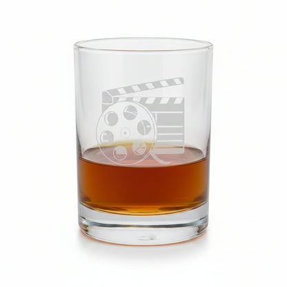 A clear glass rocks tumbler with an etched film clapperboard and reel design, partially filled with amber whiskey, set against a white background.