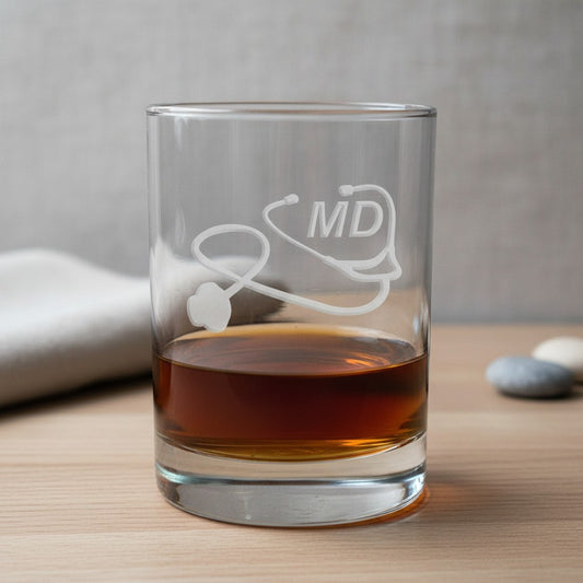 A clear glass rocks tumbler with an etched stethoscope and 'MD' design, partially filled with amber whiskey, set on a light wood surface with a blurred neutral background.