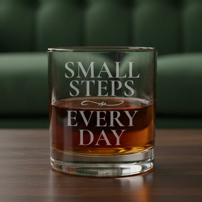 A clear lowball-style drinking glass with etched words reading 'SMALL STEPS EVERY DAY,' filled with an amber-red liquid, shown on a wood surface with green couch in the background.