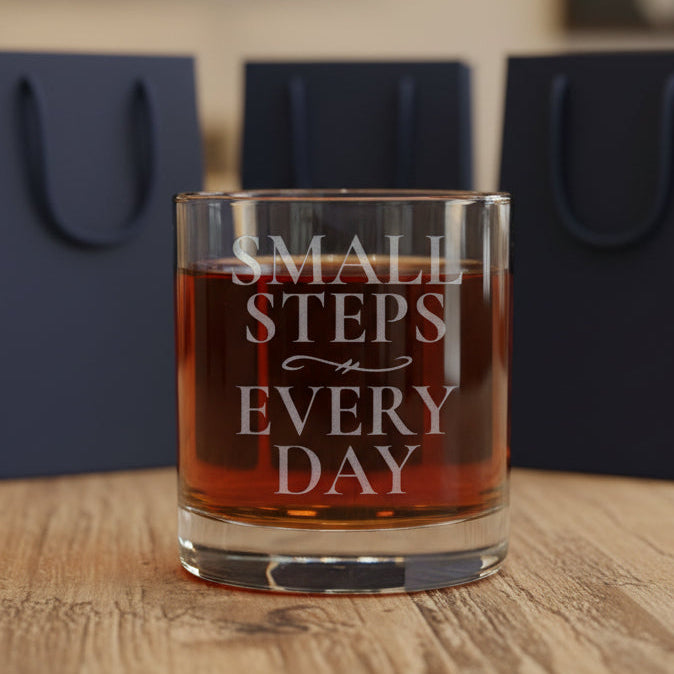 A clear lowball-style drinking glass with etched words reading 'SMALL STEPS EVERY DAY,' filled with an amber-red liquid, shown on a wood surface with gift bags in the background.