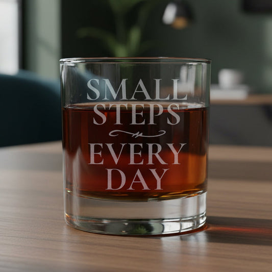 A clear lowball-style drinking glass with etched words reading 'SMALL STEPS EVERY DAY,' filled with an amber-red liquid, shown on a wood surface with a living room in the background.