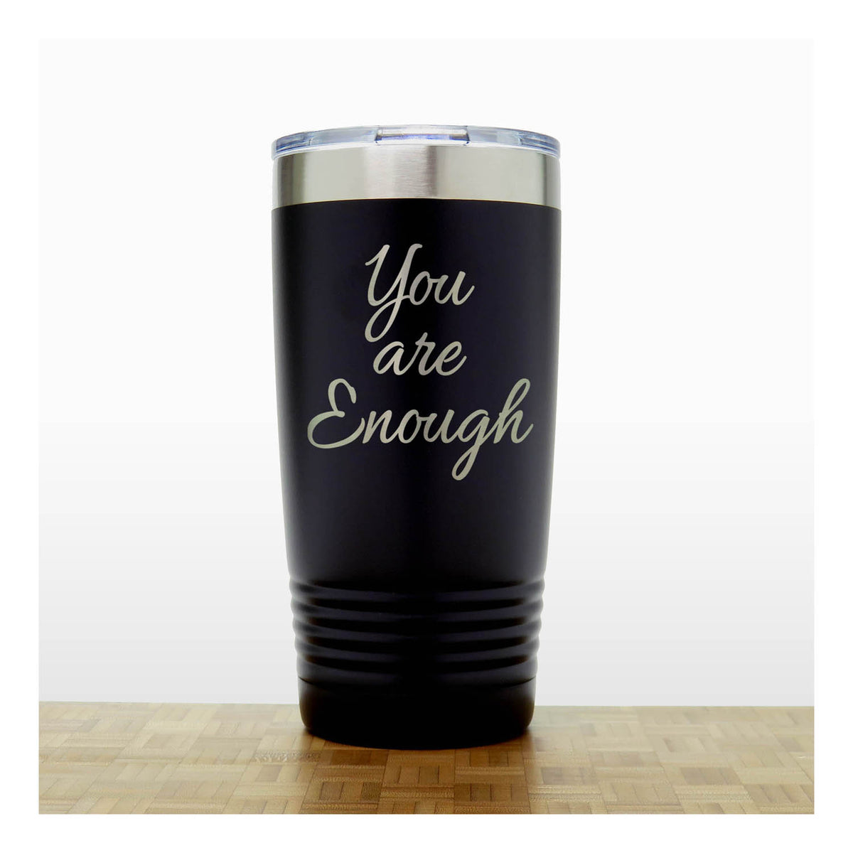 A black insulated travel tumbler with a clear lid, with laser engraved  phrase 'You are Enough' , standing on a wooden surface against a white background.