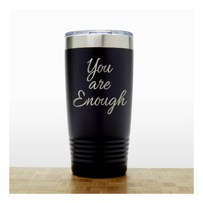 A black insulated travel tumbler with a clear lid, with laser engraved  phrase 'You are Enough' , standing on a wooden surface against a white background.