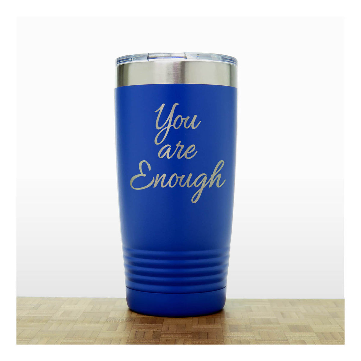 A royal blue insulated travel tumbler with a stainless steel rim and a clear lid, with laser engraved phrase 'You are Enough' . The tumbler is shown standing on a light wood surface against a white background.