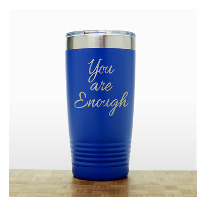 A royal blue insulated travel tumbler with a stainless steel rim and a clear lid, with laser engraved phrase 'You are Enough' . The tumbler is shown standing on a light wood surface against a white background.