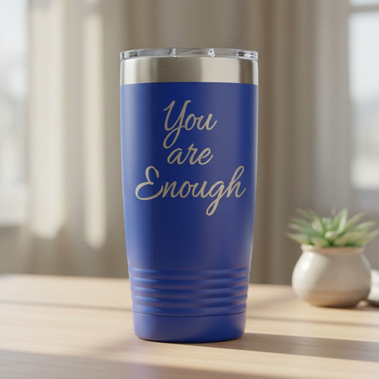A royal blue insulated tumbler with a clear lid sits on a light wooden table. The front of the tumbler features the phrase 'You are Enough' laser engraved in script font.