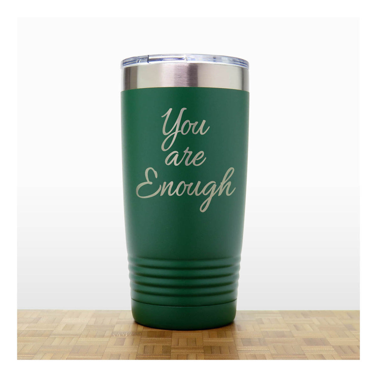 A dark green insulated tumbler with a clear lid and the words 'You are Enough' laser engraved on the front, standing on a wooden surface against a white background.