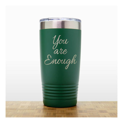 A dark green insulated tumbler with a clear lid and the words 'You are Enough' laser engraved on the front, standing on a wooden surface against a white background.