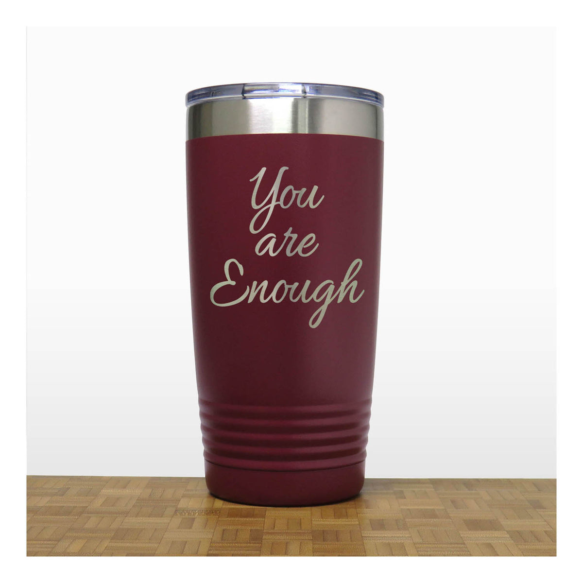 A dark red insulated travel tumbler with a stainless steel lid, with laser engraved text 'You are Enough' in a light metallic script. The tumbler is shown standing on a wooden surface against a white background.