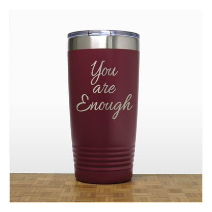 A dark red insulated travel tumbler with a stainless steel lid, with laser engraved text 'You are Enough' in a light metallic script. The tumbler is shown standing on a wooden surface against a white background.