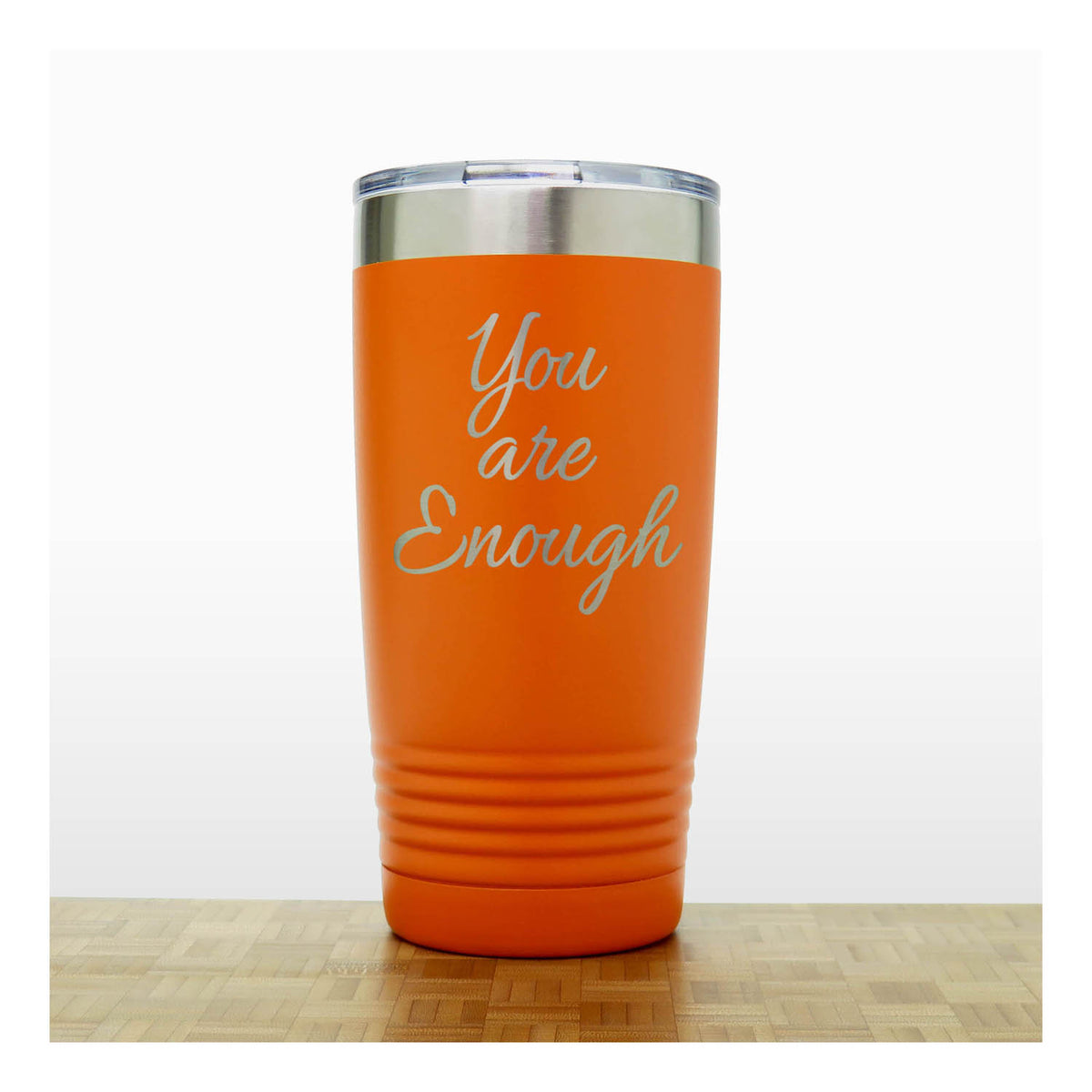 An orange insulated travel tumbler with a silver stainless steel lid and the words 'You are Enough' laser engraved on the front, standing on a light wood surface against a white background.