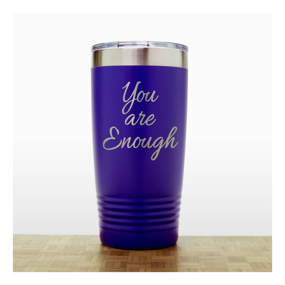 A purple insulated travel tumbler with a clear lid and silver rim, laser engraved  text 'You are Enough',  standing on a light wood surface against a white background.