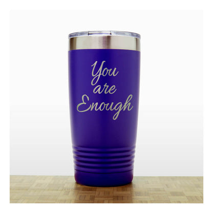 A purple insulated travel tumbler with a clear lid and silver rim, laser engraved  text 'You are Enough',  standing on a light wood surface against a white background.