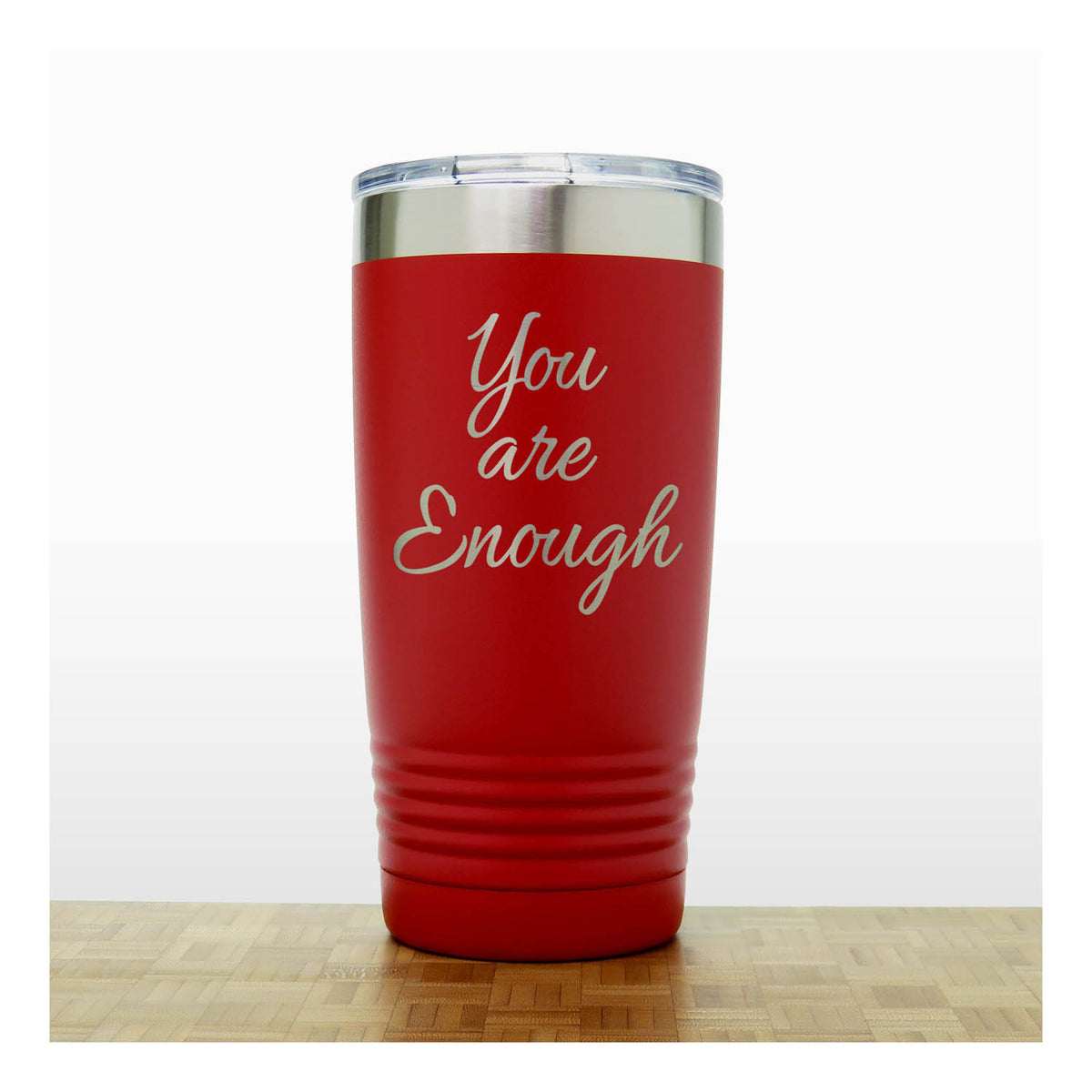 A red insulated tumbler with a clear lid and silver metal rim, laser engraved  phrase 'You are Enough'  on the front, shown standing on a light wood surface against a white background.