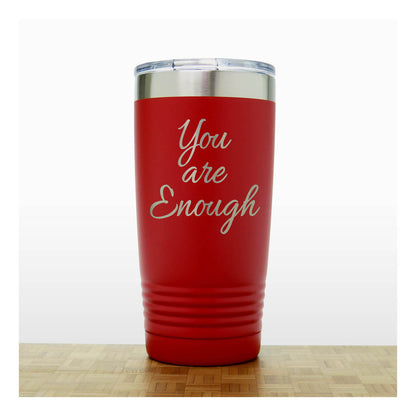 A red insulated tumbler with a clear lid and silver metal rim, laser engraved  phrase 'You are Enough'  on the front, shown standing on a light wood surface against a white background.
