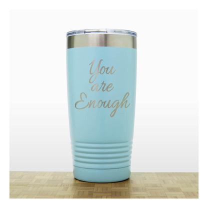 A light blue insulated travel tumbler with a clear lid and a metallic gold laser-engraved quote reading 'You are Enough' on the front, standing on a wooden surface against a white background.