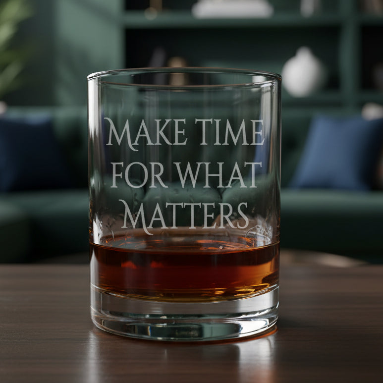 A clear rocks glass with the phrase 'MAKE TIME FOR WHAT MATTERS' etched on the front, partially filled with amber whiskey, set on a wood surface against a living room background.