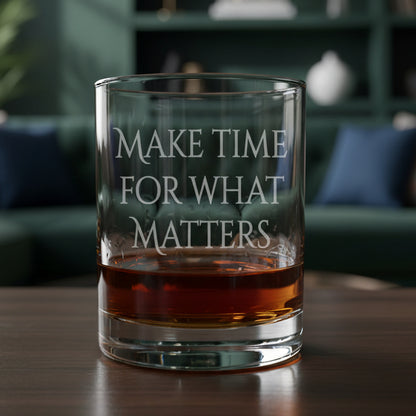 A clear rocks glass with the phrase 'MAKE TIME FOR WHAT MATTERS' etched on the front, partially filled with amber whiskey, set on a wood surface against a living room background.