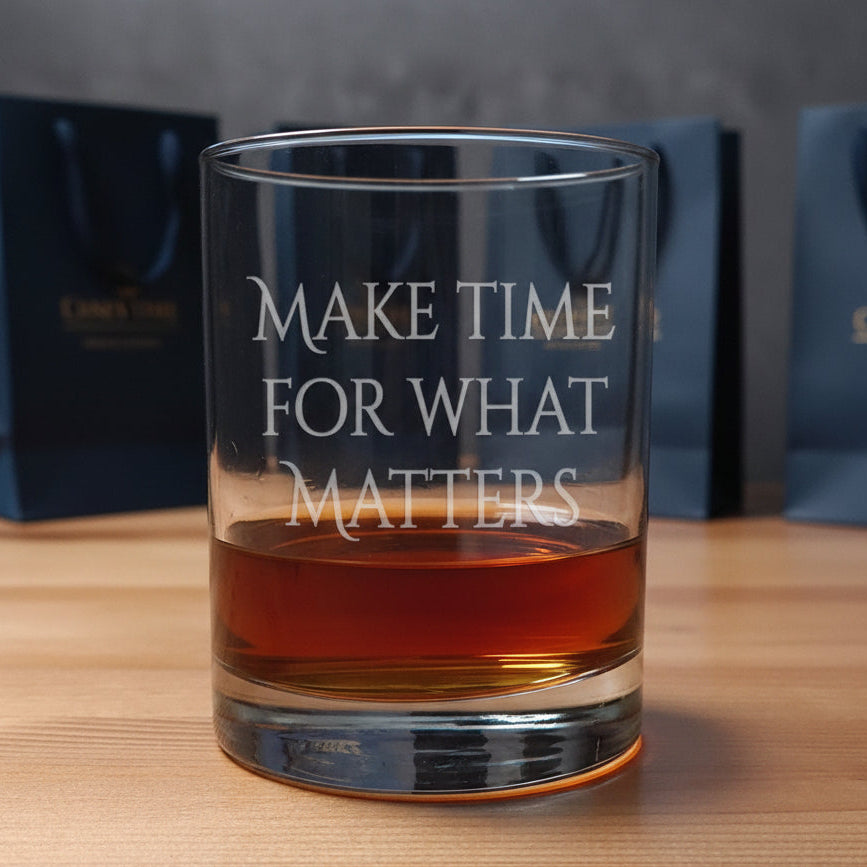 A clear rocks glass with the phrase 'MAKE TIME FOR WHAT MATTERS' etched on the front, partially filled with amber whiskey, set on a wood surface 
